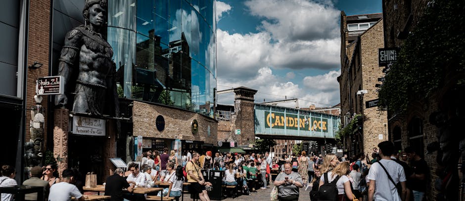A busy street scene in Camden Town NW1 with numerous pedestrians walking and sitting at outdoor tables of cafes and restaurants under a partly cloudy sky. In the background, a loading area features a man with van Camden actively engaged in a home relocation process, loading or unloading cardboard boxes, wrapped furniture, and household items into or from a white van. The area near the pavement displays various materials such as cardboard boxes, plastic wrap, and blankets used for packing and protecting furniture during transportation. The street is lined with brick buildings, some with outdoor signage, and the presence of a large glass-fronted building with a statue visible on the left side. The scene captures a typical urban environment where a professional removals company is assisting with furniture transport, packing, and moving logistics amidst a lively community atmosphere.