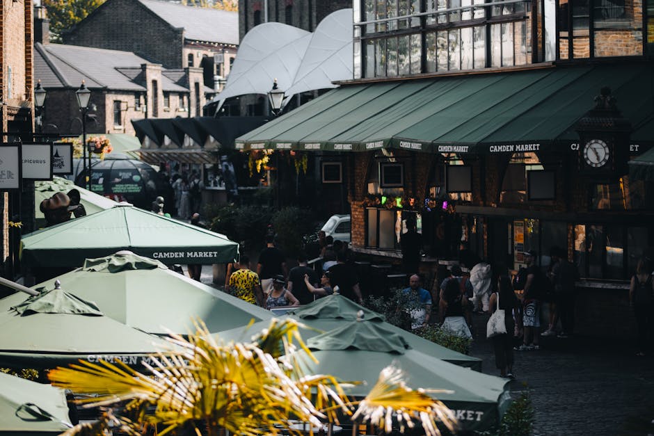 A busy street scene at Camden Market with outdoor seating areas covered by green umbrellas displaying 'Camden Market' branding. In the background, there are brick buildings with large windows, and a modern, curved white architectural structure. The sidewalk is crowded with pedestrians, some walking and others standing in small groups, engaged in conversation. To the left, black vans and cars are parked along the street, and some of the vehicles are visible behind the umbrellas. The scene suggests an active marketplace environment with a mix of outdoor leisure and shopping activities, typical of a central London relocation process involving home or commercial furniture transport, packing, or moving logistics in narrow street conditions. The lighting indicates natural daylight, enhancing the visibility of various objects and the lively atmosphere typical of Camden's outdoor market area, which [MAN WITH VAN CAMDEN] would facilitate for household or business removals. 