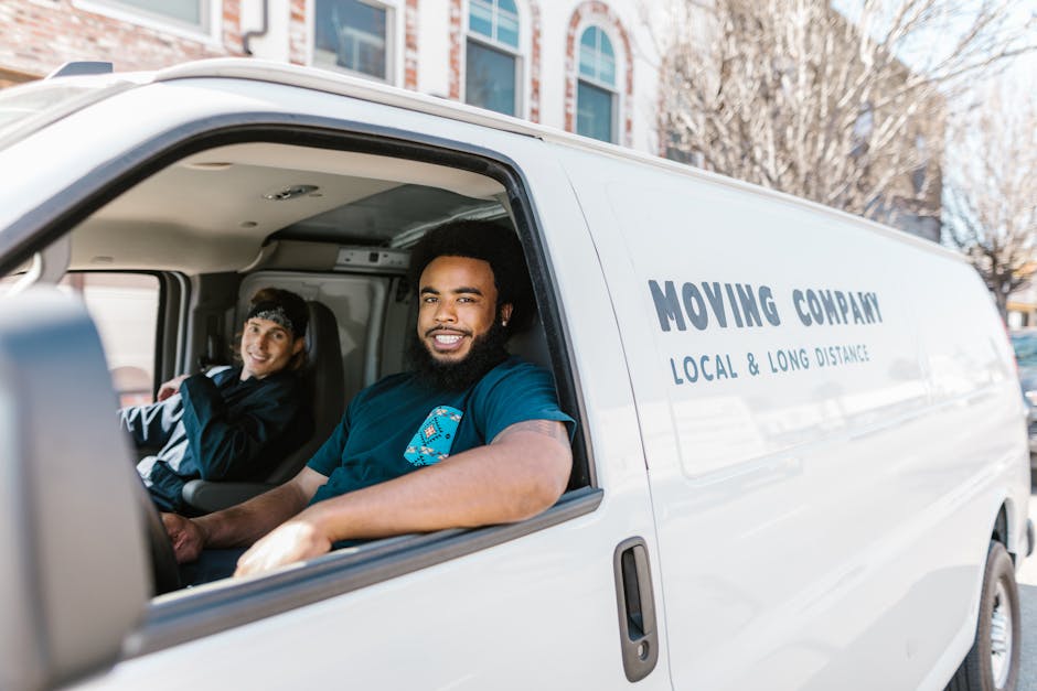 Inside the cab of a white moving van labeled 'MOVING COMPANY LOCAL & LONG DISTANCE,' two men are seated; one in the driver’s seat and the other in the passenger seat, both smiling at the camera. The driver, an African-style man with curly hair and a beard, is wearing a black t-shirt with a blue logo, and the passenger, a young man with light skin and dark hair, is wearing a black jacket with a grey cap. The van is parked outdoors on a paved street during daylight, with residential buildings and leafless trees visible in the background. The interior of the van shows its ceiling with built-in lighting and some packing materials, indicating readiness for home relocation or furniture transport. Man with Van Camden specializes in house removals, including packing and moving logistics, as part of their professional services for estate flat relocations in Camden, NW5, and surrounding areas.