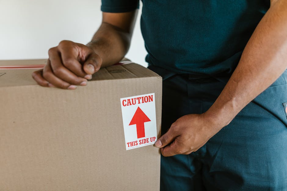 Close-up of a person wearing a dark green shirt and blue work trousers, holding a large cardboard box during a home relocation process. The individual is attaching a red and white caution label with an upward arrow and the text 'CAUTION THIS SIDE UP' to the side of the box. The box appears to be part of packed belongings, potentially for an estate flat move in Kentish Town, with the focus on the careful handling and packing process. The background shows an indoor area with a plain, light-colored wall, and the lighting is natural and even, emphasizing the importance of proper packing and marking for furniture transport and loading during removals. The image illustrates key elements of packing and moving logistics managed by companies like Man with Van Camden, ensuring safe handling of items during the loading process.