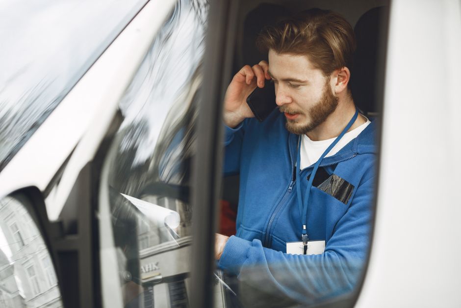 A male mover with a beard, dressed in a red and navy jacket, black cargo trousers, and a blue beanie, stands in front of an open van loaded with neatly stacked cardboard boxes. The boxes are sealed wi
